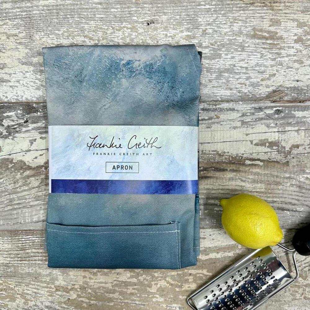 Frankie Creith DUNLUCE Apron folded with blue design showing castle print on cotton fabric with lemon and grater on wooden background